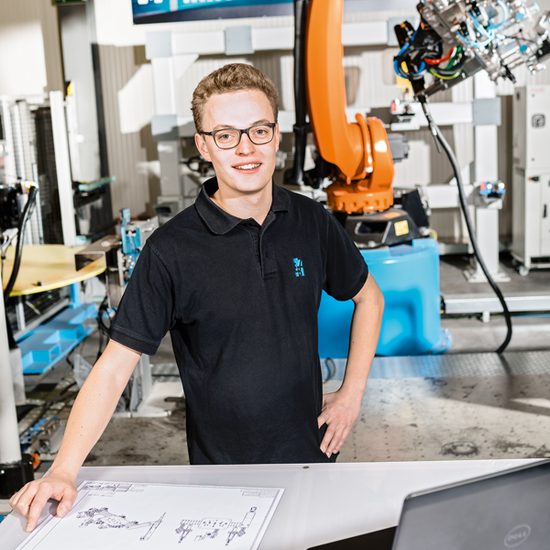 Ein angehender Mechatroniker arbeitet anhand einer technischen Zeichnung. Im Hintergrund ist ein Industrieroboter zu sehen.