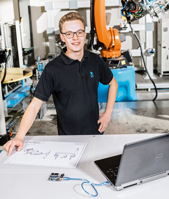 Ein angehender Mechatroniker arbeitet anhand einer technischen Zeichnung. Im Hintergrund ist ein Industrieroboter zu sehen.